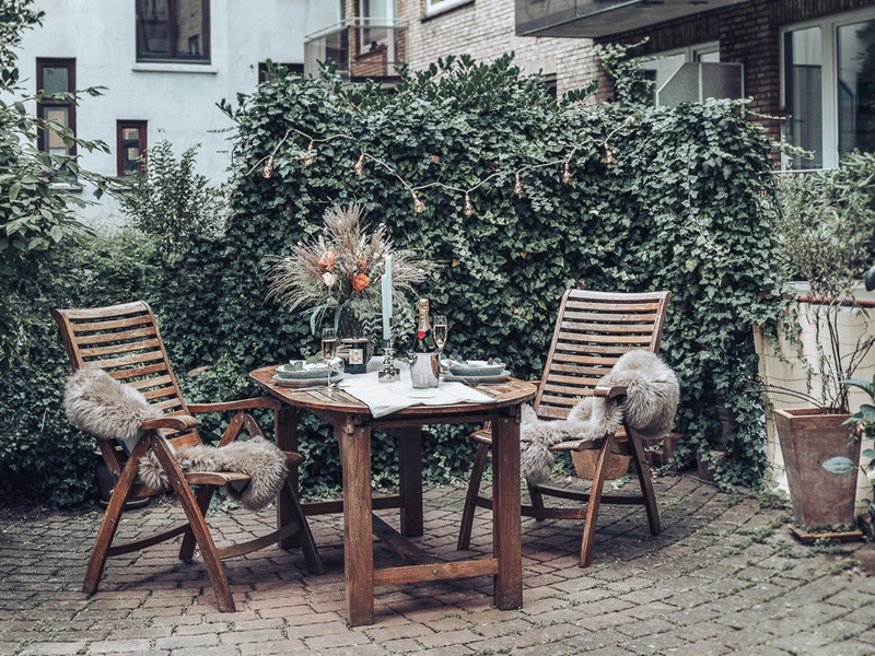 Holztisch mit zwei Stühlen und Dekoration im gemütlichen Garten, umgeben von grüner Hecke.