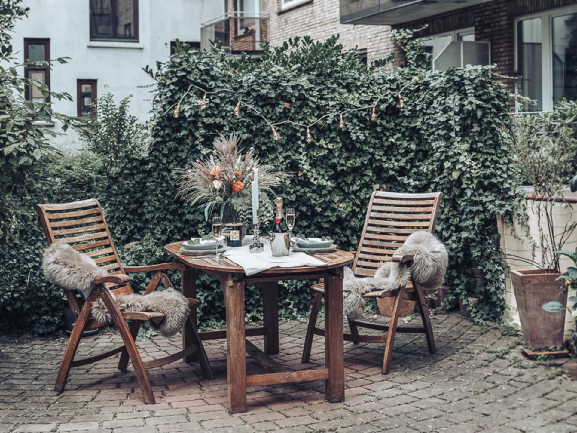 Holztisch mit zwei Stühlen und Dekoration im gemütlichen Garten, umgeben von grüner Hecke.