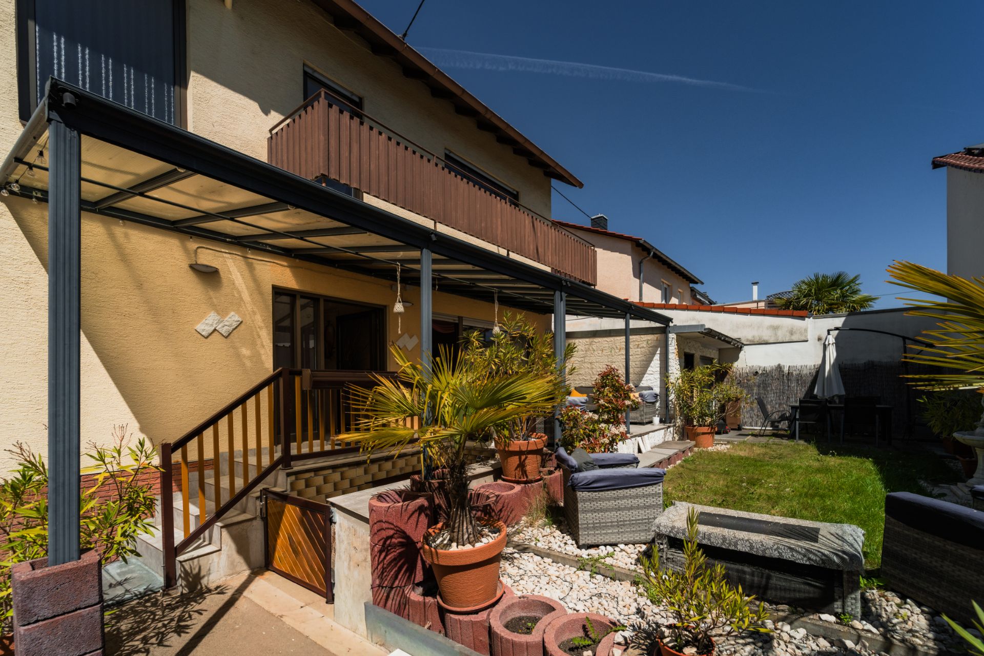 Terrasse mit Überdachung, Gartenmöbeln, Pflanzen in Töpfen und kleinem Rasen im Hinterhof eines Hauses.