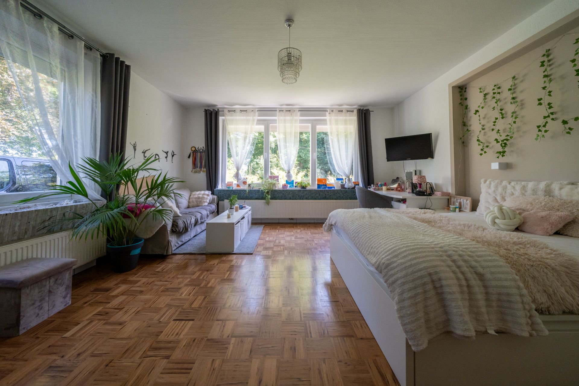 Helles Schlafzimmer mit großem Fenster, Bett, Sofa, Schreibtisch und Pflanzen.