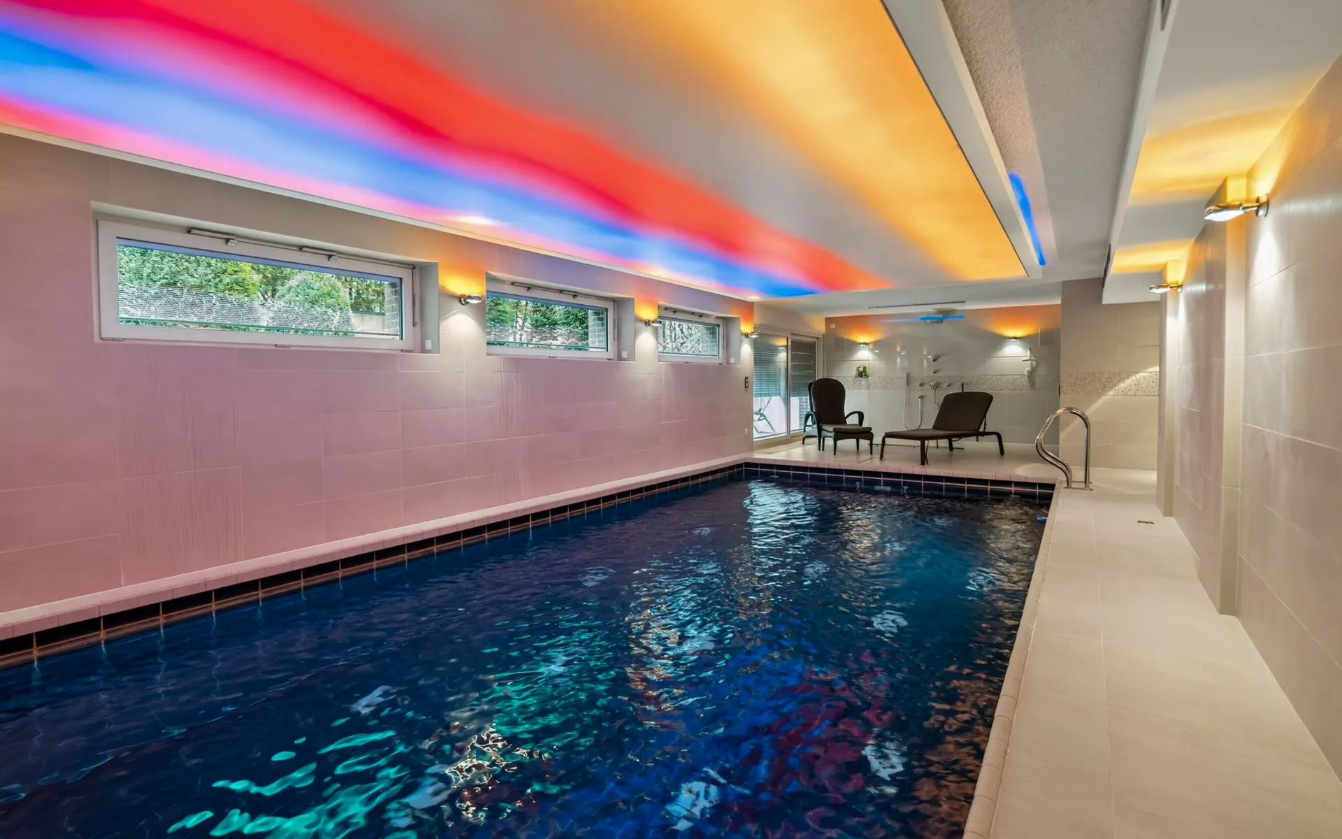 Heller Innenpool mit buntem Deckenlicht, seitlichen Fenstern und zwei Liegestühlen am Rand.