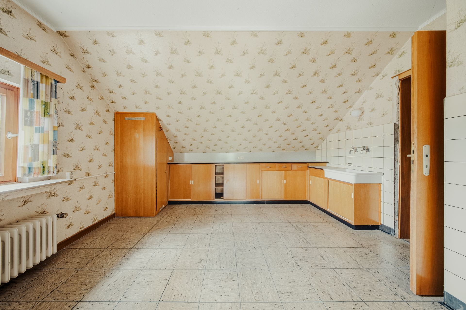 Leere Küche mit Schrägdach, Holzmöbeln, Fenster mit Vorhang und Heizkörper.