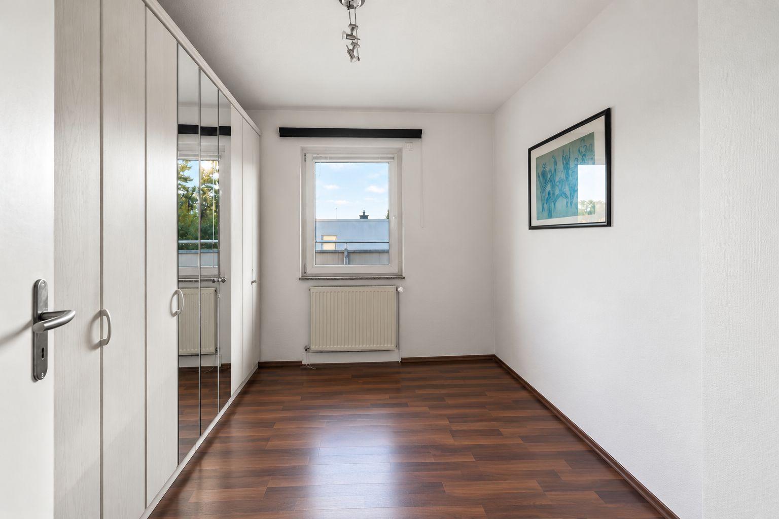 Leeres Zimmer mit Holzboden, weißem Kleiderschrank, Fenster und Wandbild.
