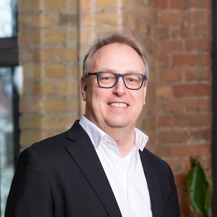 Mann mit Brille im Anzug steht vor einer Backsteinwand und einer Pflanze.