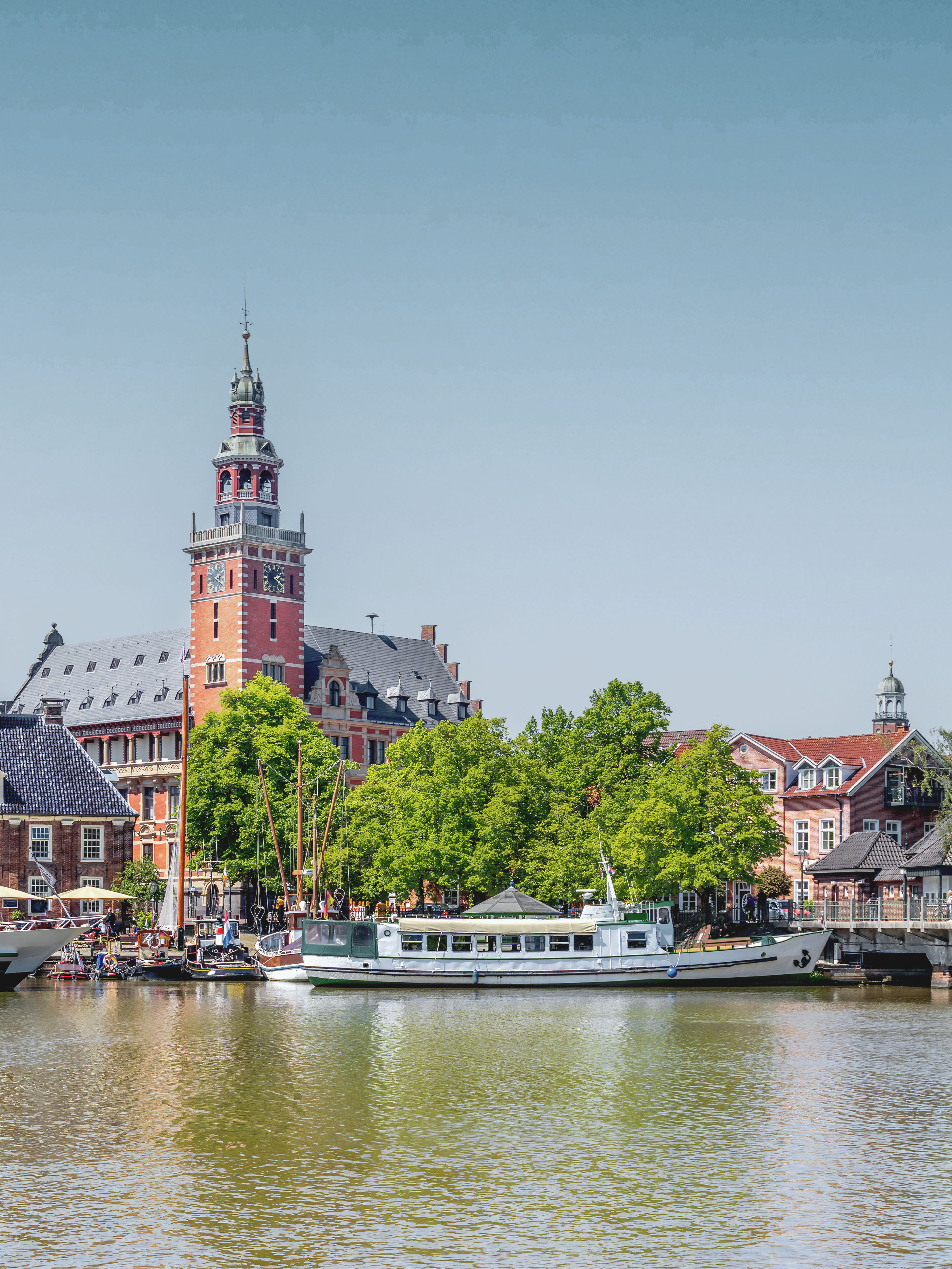 Backsteingebäude mit Uhrturm am Wasser, Boote und grüne Bäume im Vordergrund.
