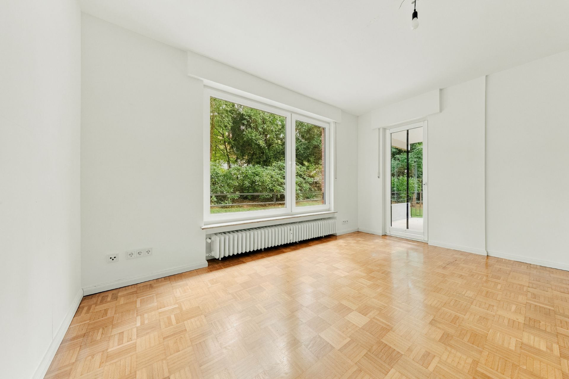 Helles, leeres Zimmer mit Parkettboden, großem Fenster und Tür zur Terrasse, Blick ins Grüne.