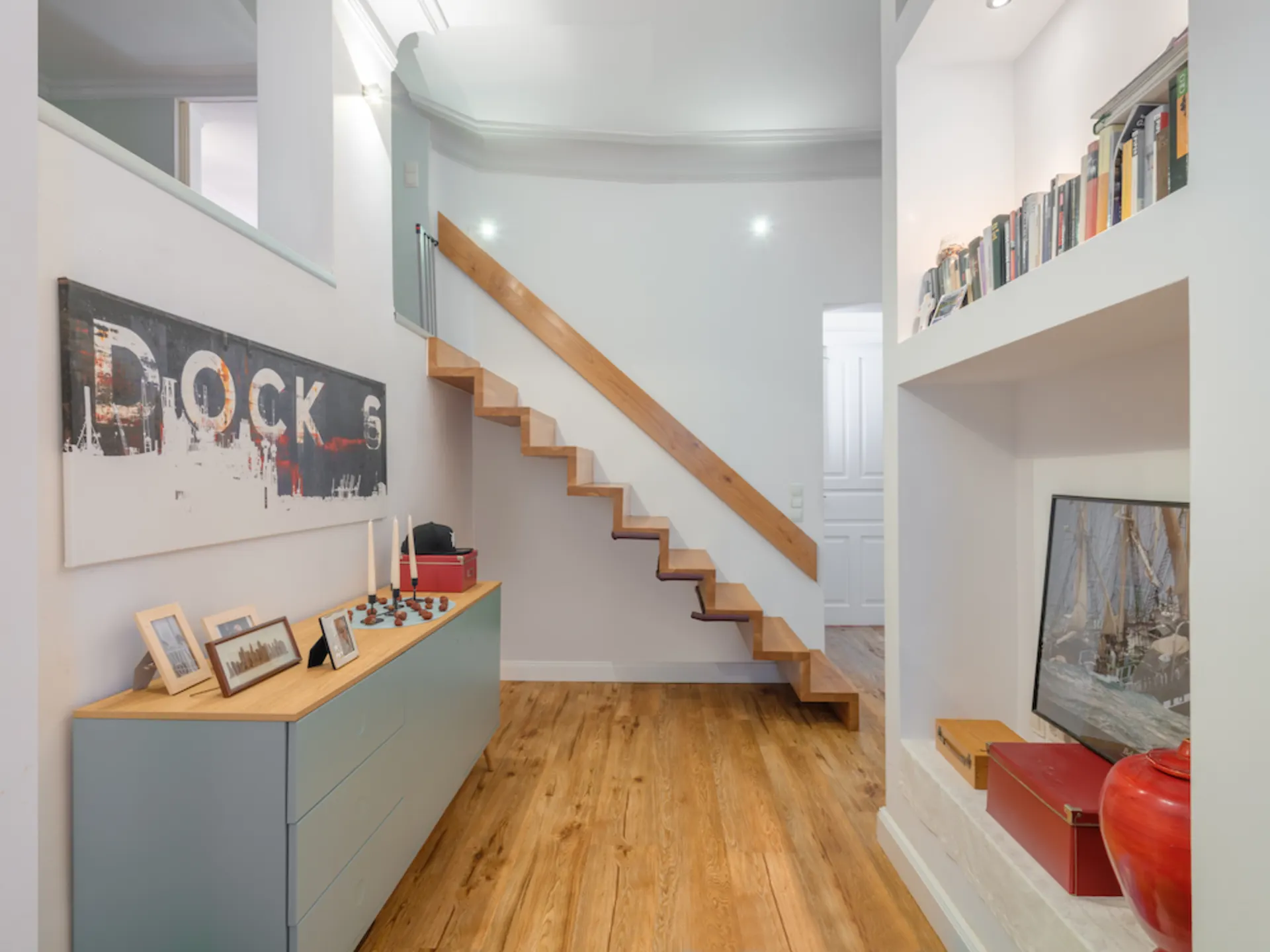 Moderner Flur mit Holztreppe, Kommode, Bildern und Regal mit Büchern.