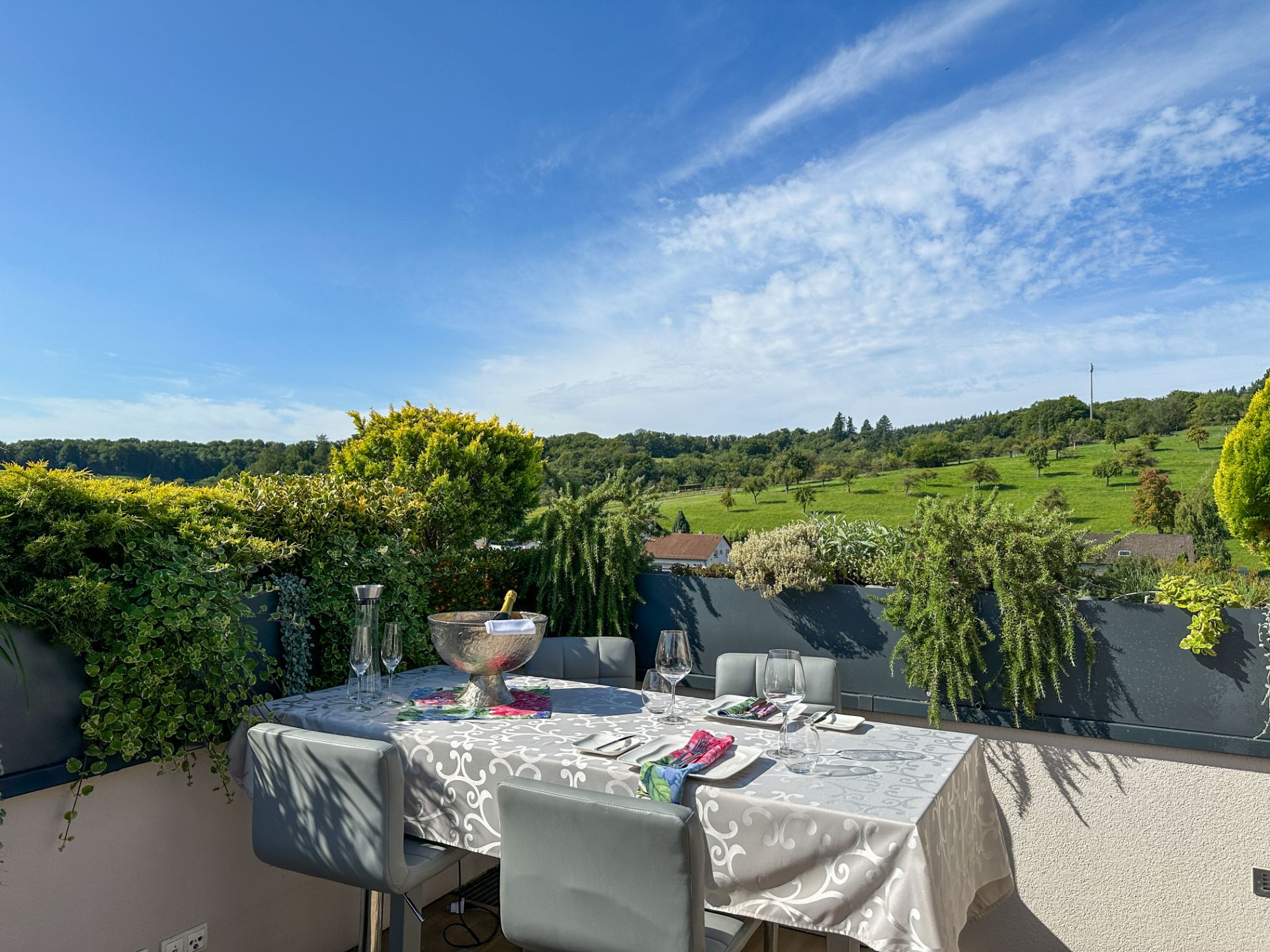 Gedeckter Tisch auf einer Terrasse mit Blick auf grüne Hügel und blauen Himmel.