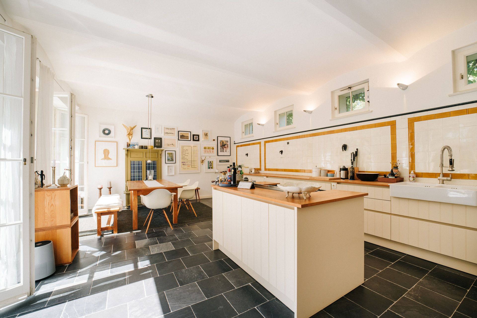 Helle Küche mit Kücheninsel, Esstisch, vielen Bildern an der Wand und großen Fenstern.