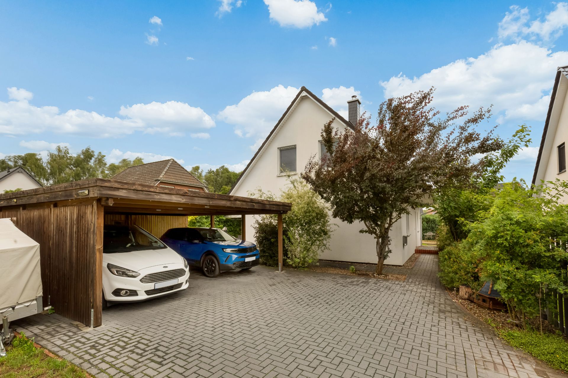 Einfamilienhaus mit Carport, zwei geparkten Autos und gepflasterter Einfahrt, daneben ein Baum.