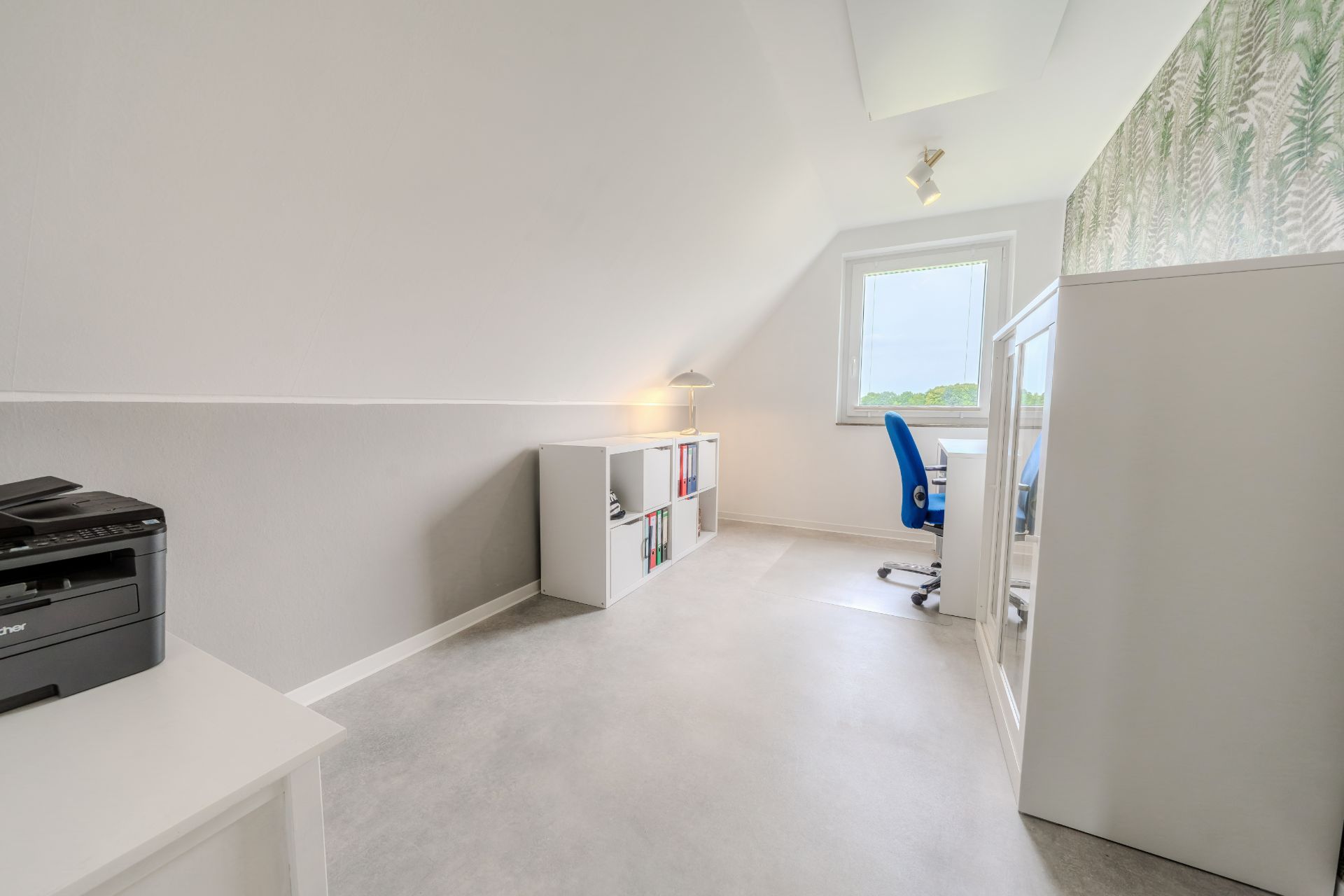 Heller Arbeitsraum mit Schreibtisch, blauem Stuhl, Regal und Drucker unter Dachschräge.