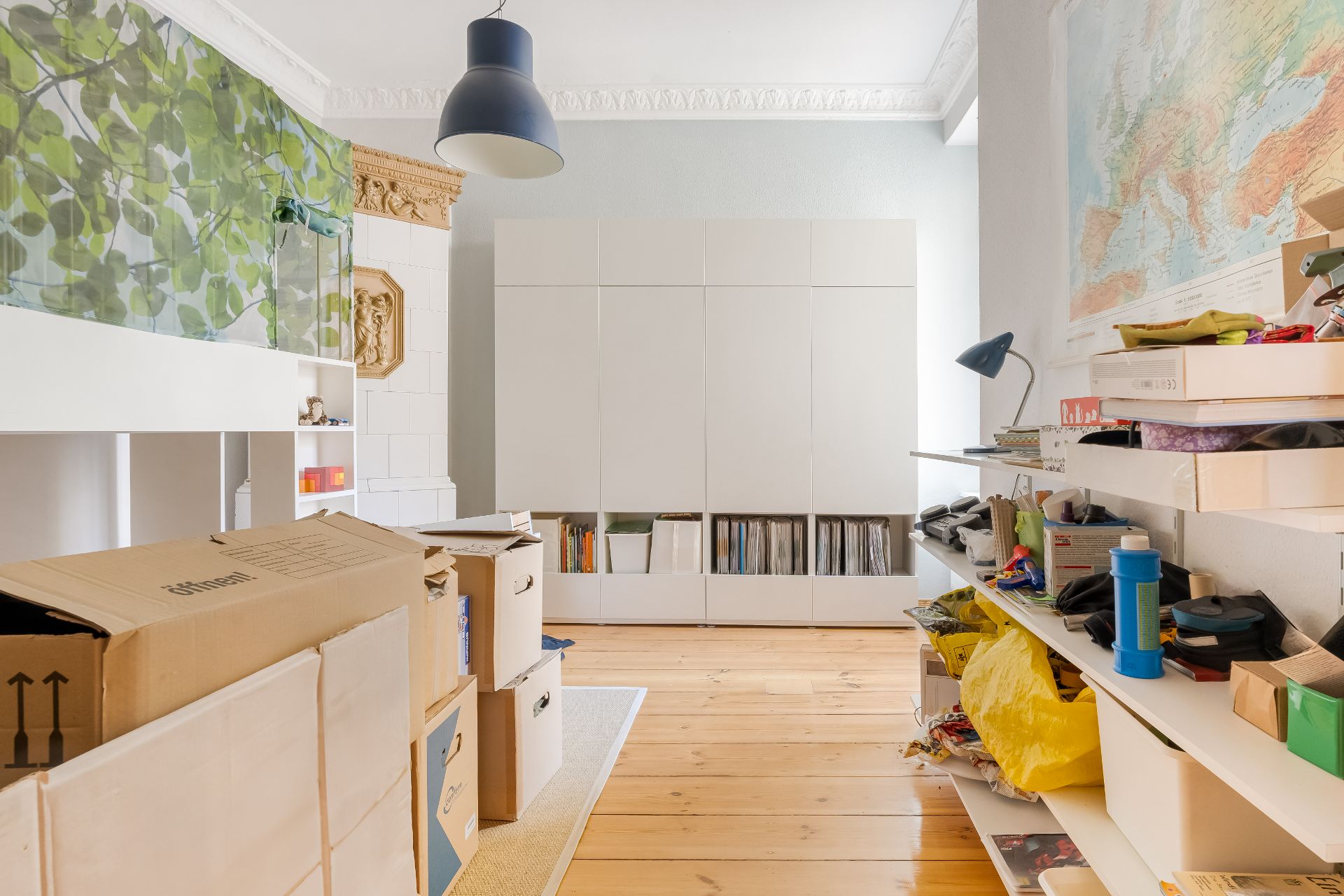 Helles Zimmer mit Kartons, weißen Regalen, Schrank und Wandkarte, Holzboden.