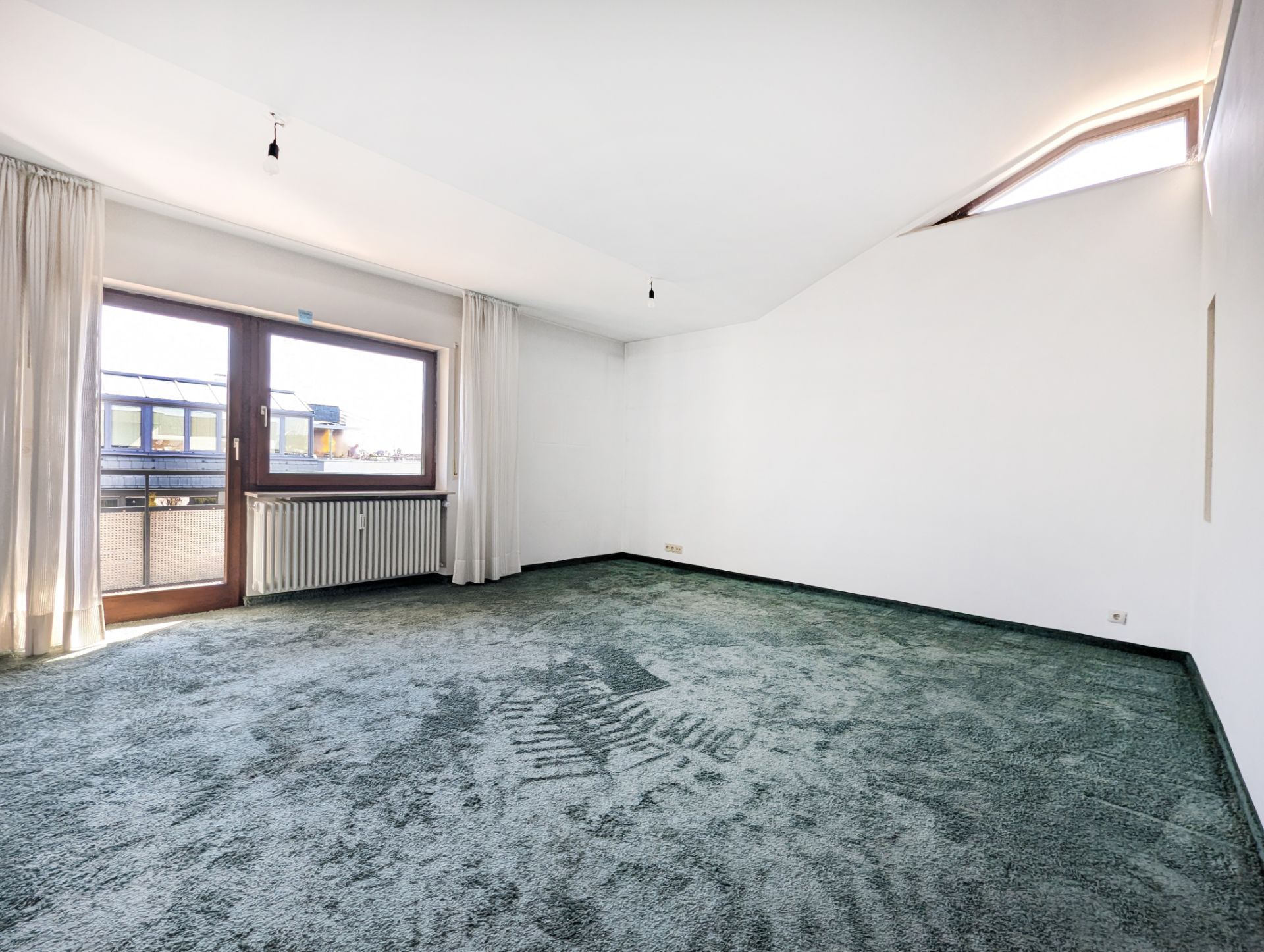 Leerer Raum mit grünem Teppich, großen Fenstern, weißen Wänden und Vorhängen.