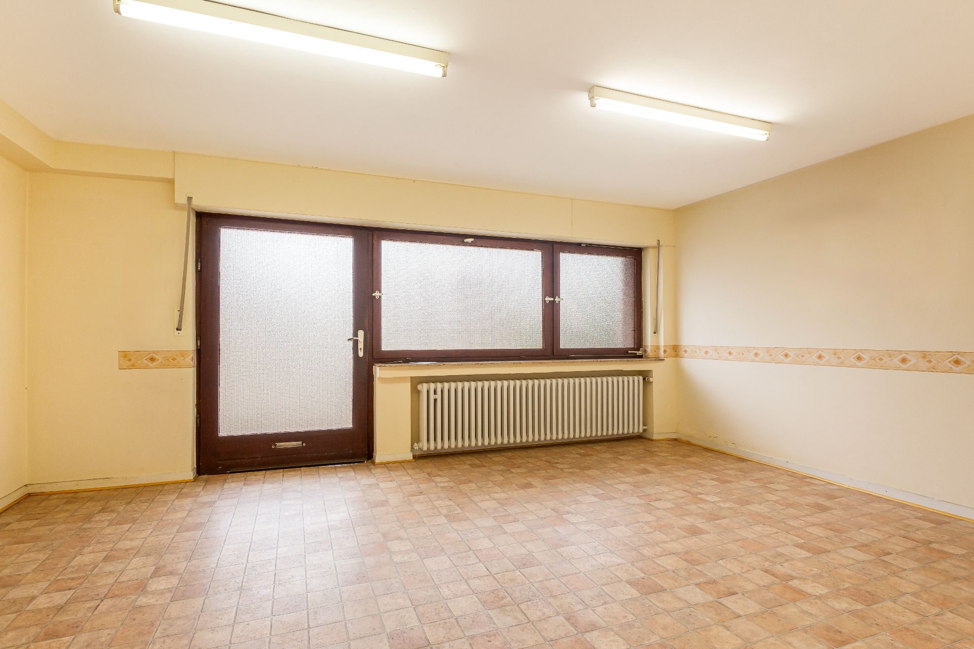Leerer Raum mit gemustertem Boden, großen Fenstern, Heizung und gelben Wänden.