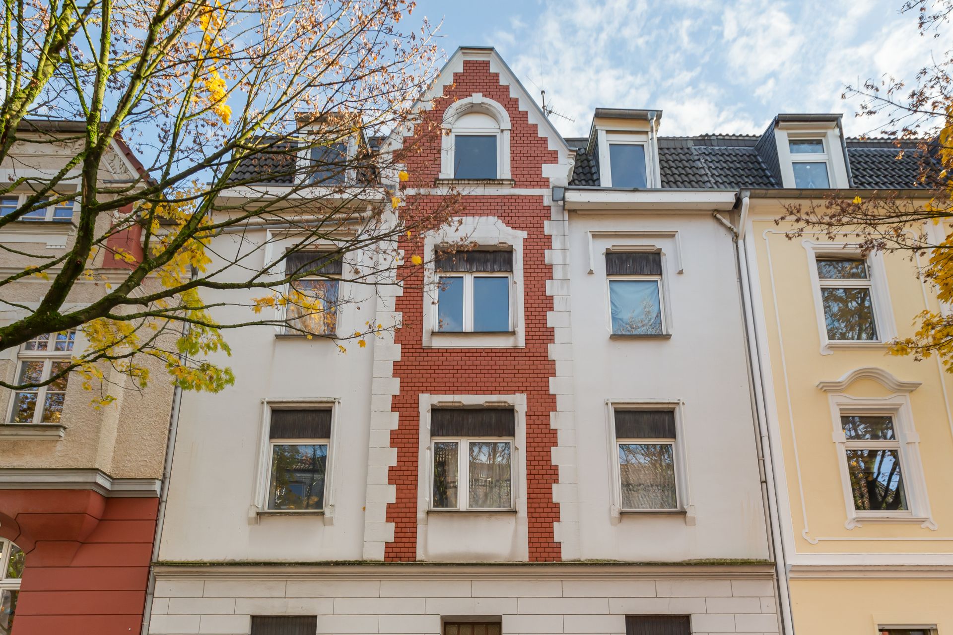 Mehrstöckiges Wohnhaus mit roter Backsteinfassade in der Mitte, umgeben von Bäumen.