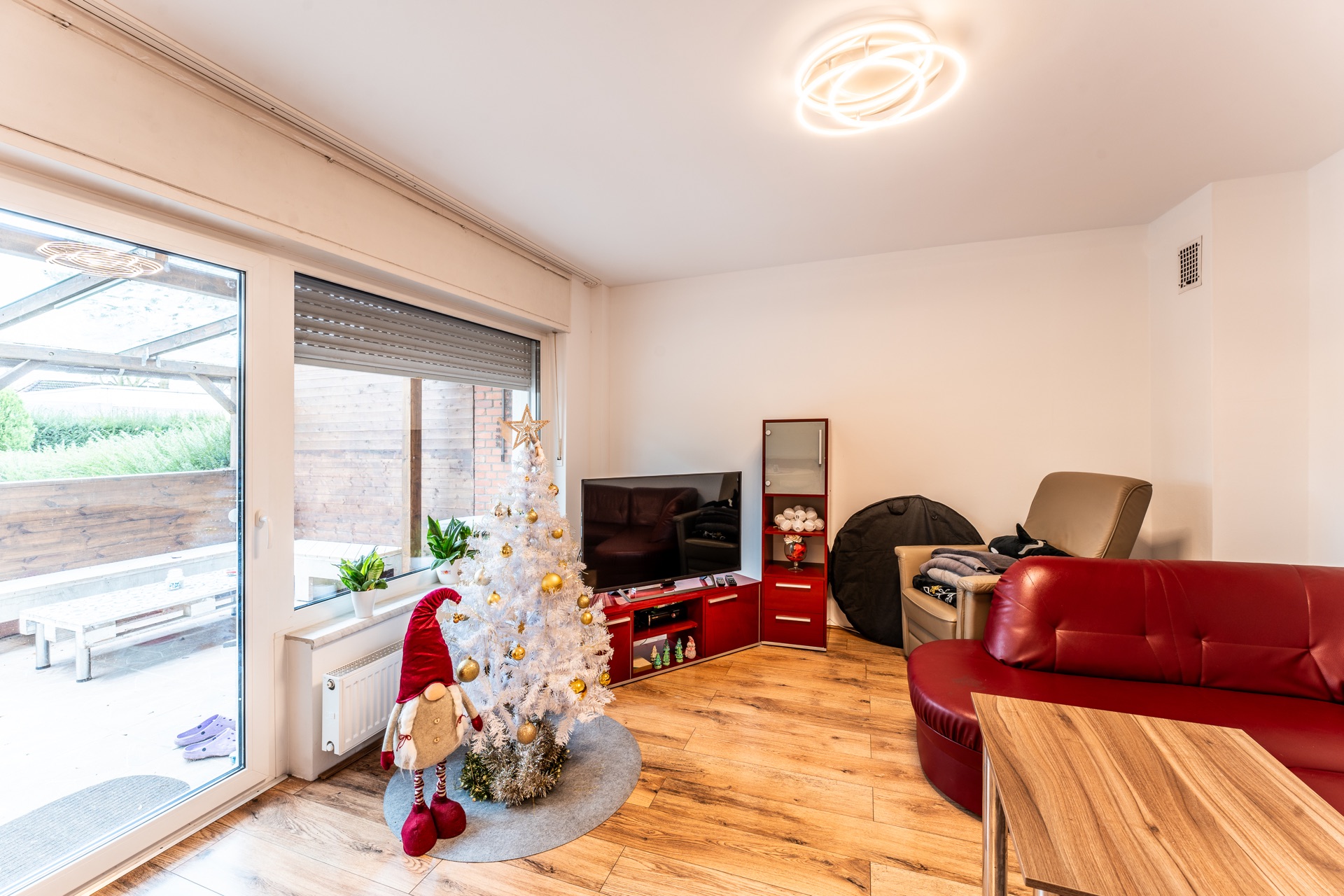 Wohnzimmer mit rotem Sofa, Fernseher, weißem Weihnachtsbaum und großen Fenstern zur Terrasse.