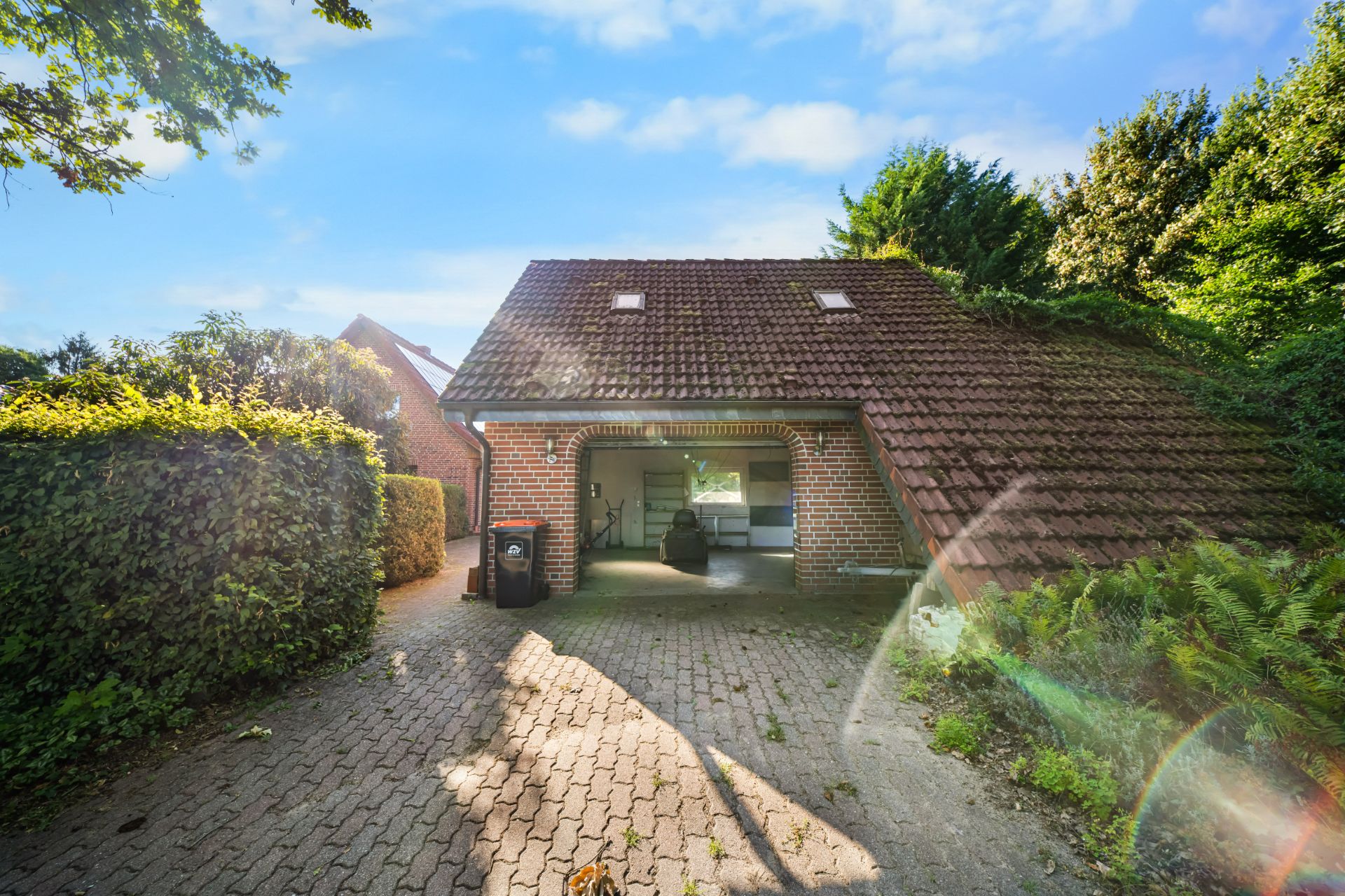 Backstein-Carport mit schrägem Dach und Einfahrt, umgeben von Hecken und Bäumen.