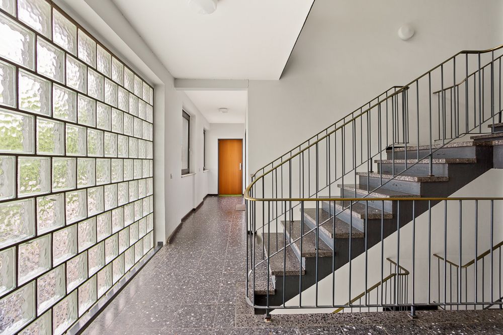 Flur mit Glaselementwand, Fenstern, Treppenhaus und Holztür am Ende.