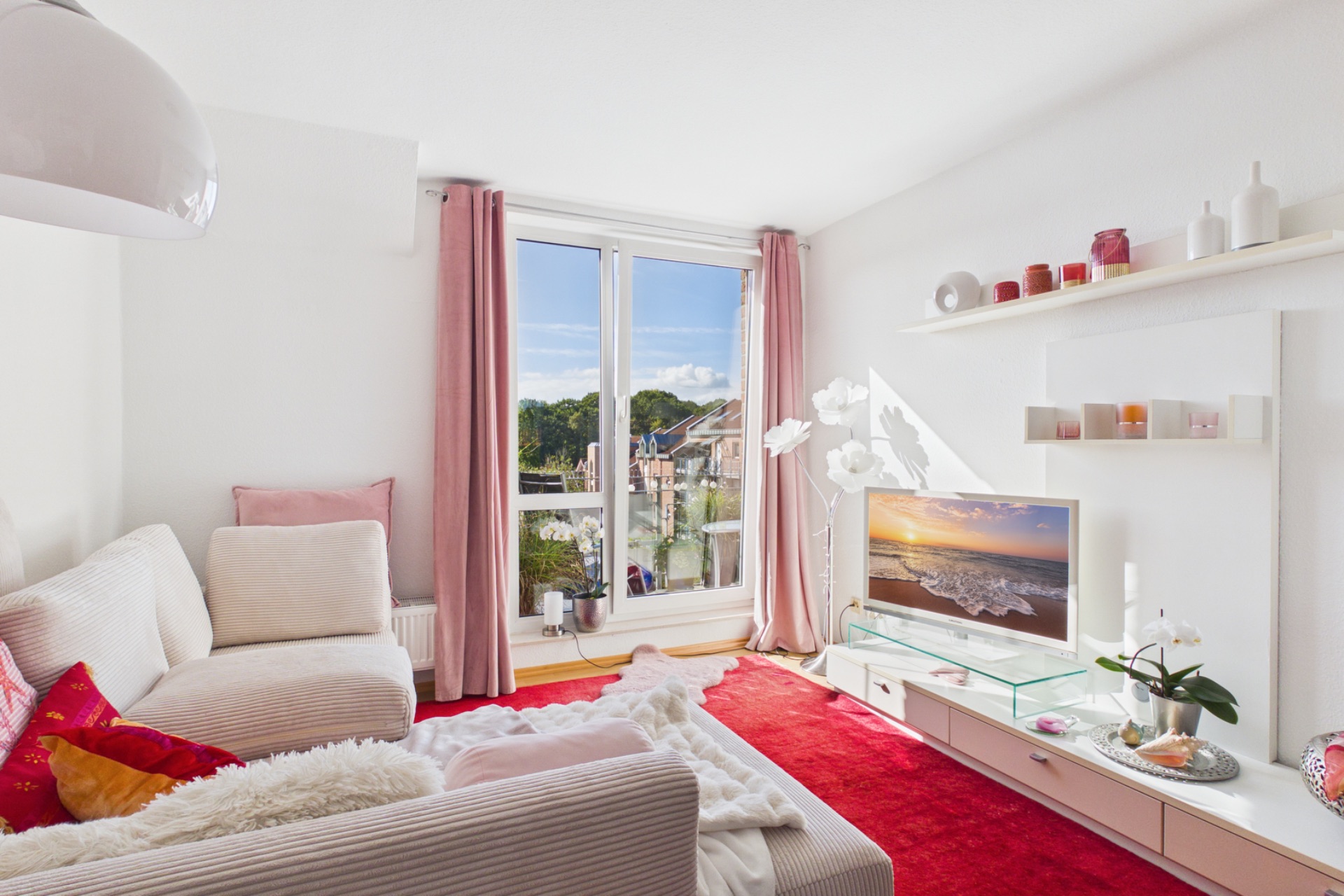 Helles Wohnzimmer mit Sofa, rotem Teppich, Fernseher und großen Fenstern mit rosa Vorhängen.
