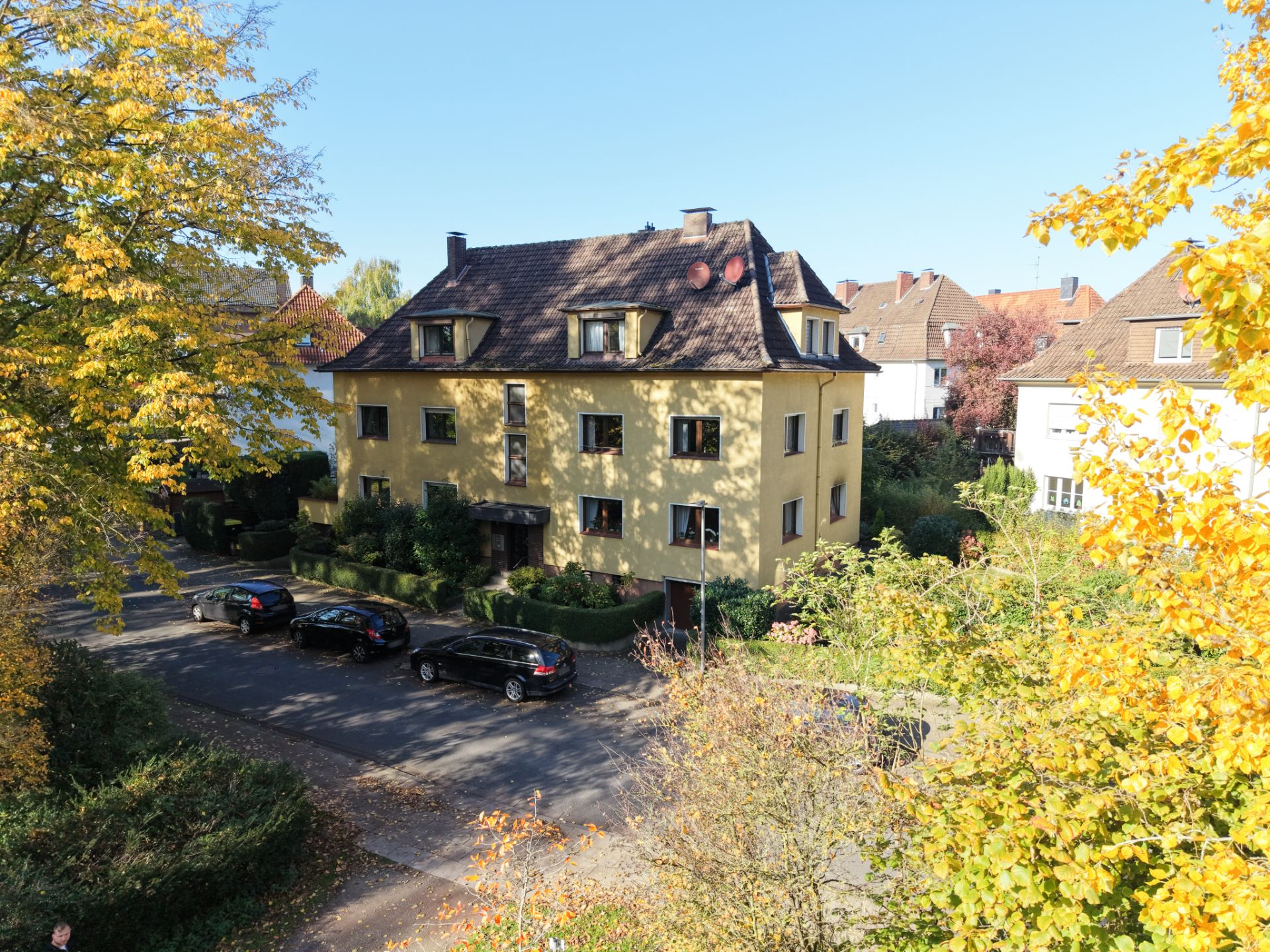 Gelbes Mehrfamilienhaus mit Garten, umgeben von Bäumen und parkenden Autos an einer ruhigen Straße.