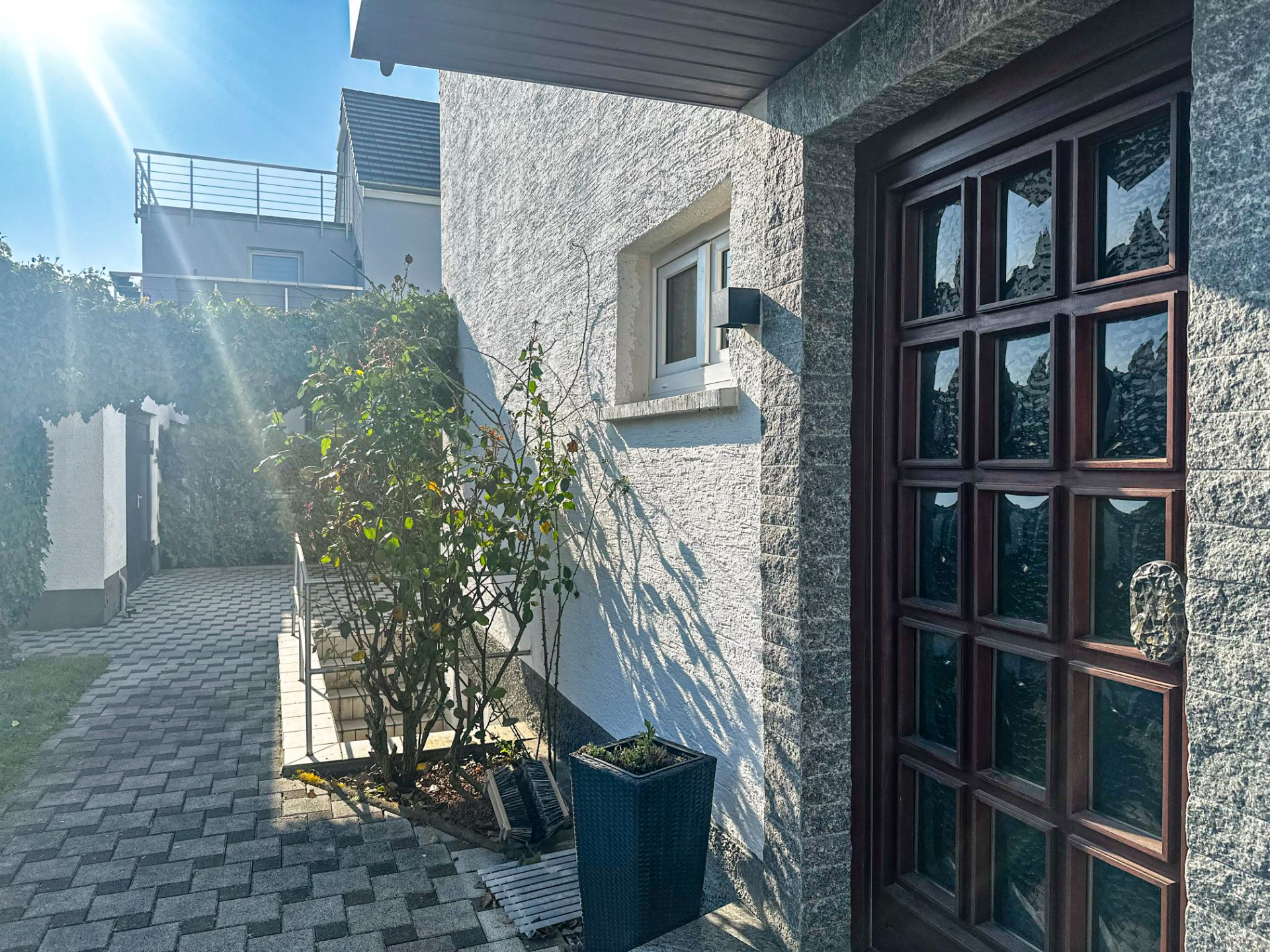 Eingangsbereich eines Hauses mit Holztür, Blumen und gepflastertem Weg bei Sonnenschein.