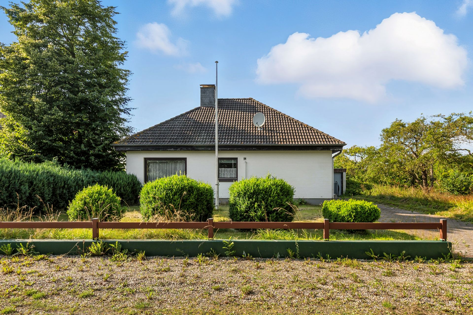 Weißes Einfamilienhaus mit dunklem Dach, umgeben von Büschen und Bäumen, im Vordergrund ein Kiesweg.