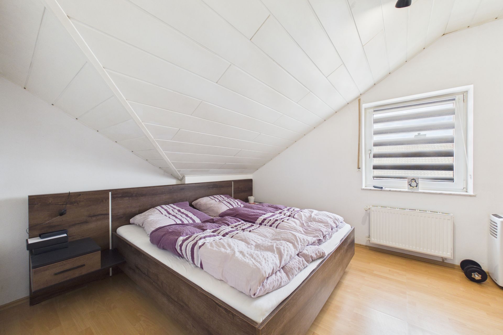 Schlafzimmer mit Doppelbett unter einer Dachschräge, Fenster mit Jalousien und Holzfußboden.