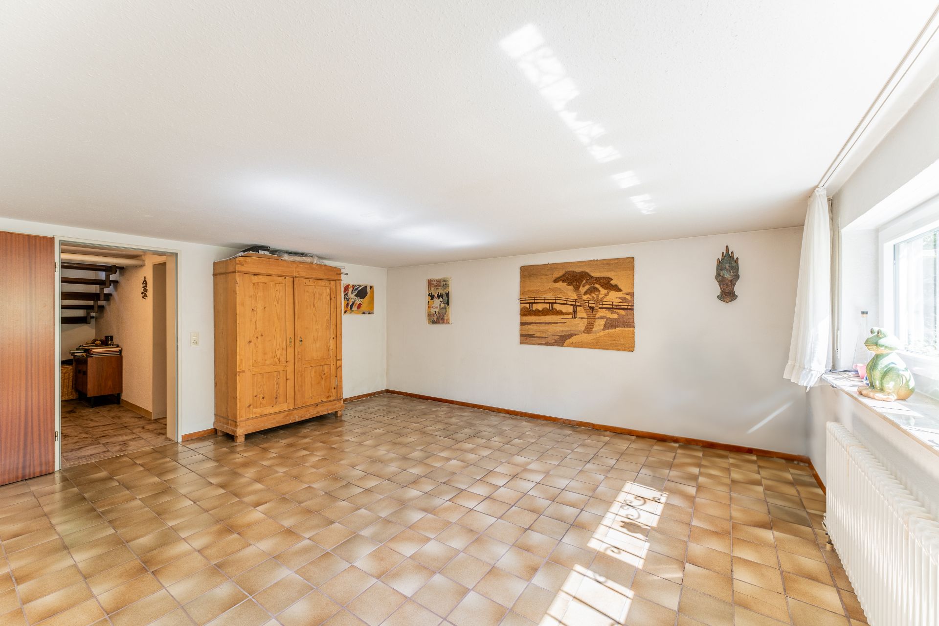 Heller Raum mit Fliesenboden, Holzschrank, Bildern an der Wand und Fenster mit Vorhang.