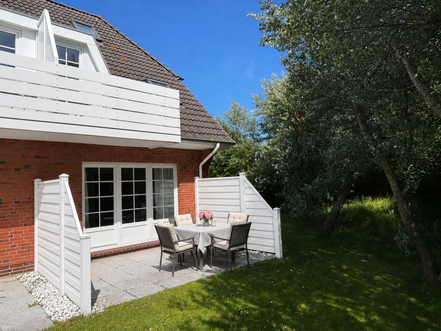 Kleine Terrasse mit Tisch und Stühlen vor einem Backsteinhaus, umgeben von Bäumen und Rasen.