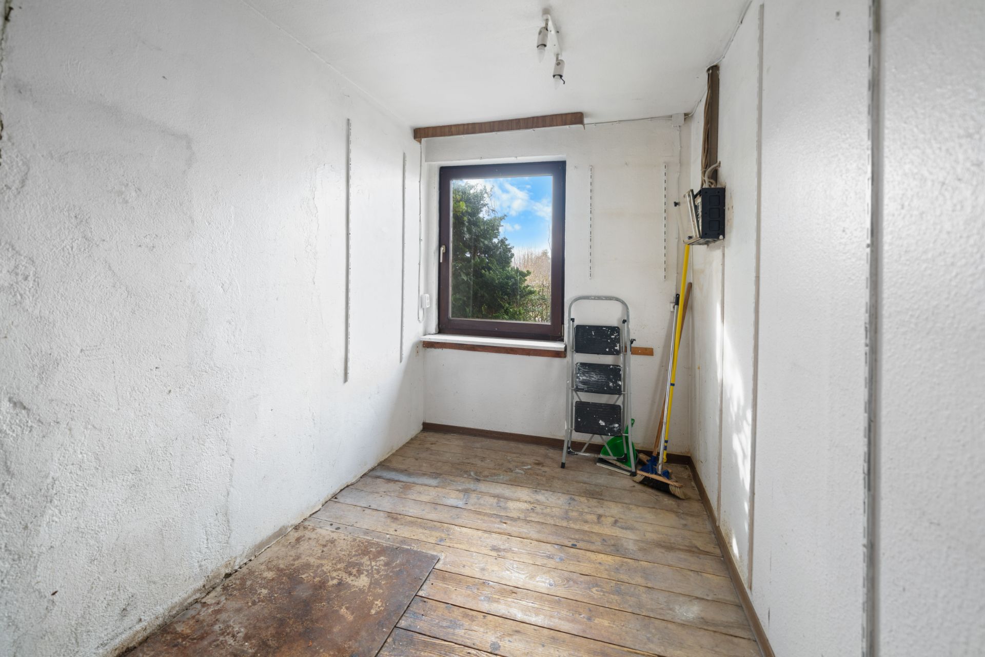 Kleiner, leerer Raum mit Holzboden, Fenster, Leiter und Putzutensilien an der Wand.