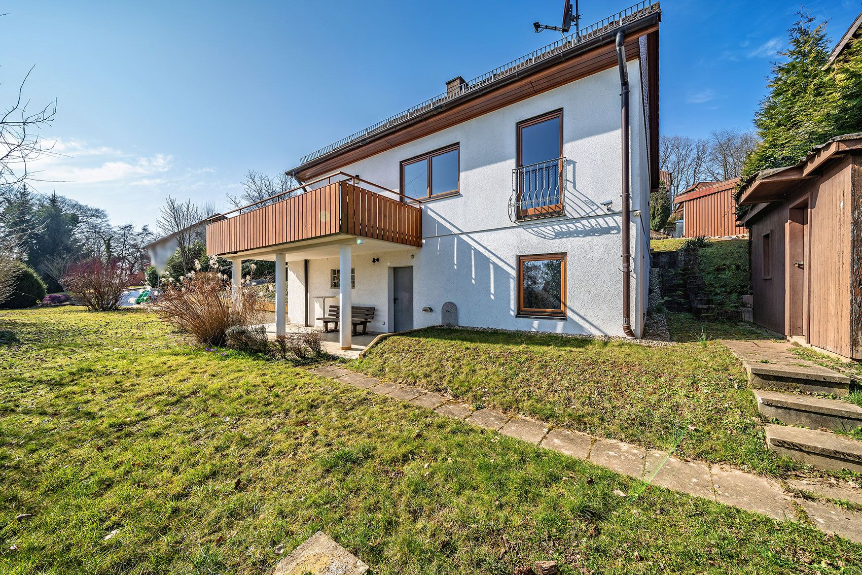 Haus mit weißer Fassade, Balkon, großen Fenstern und Garten bei sonnigem Wetter.