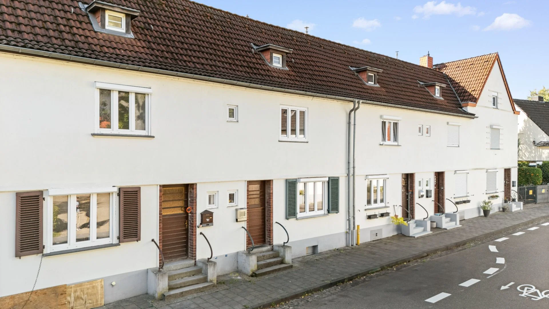 Reihenhaus mit weißer Fassade, braunen Fensterläden und mehreren Eingängen an einer ruhigen Straße.