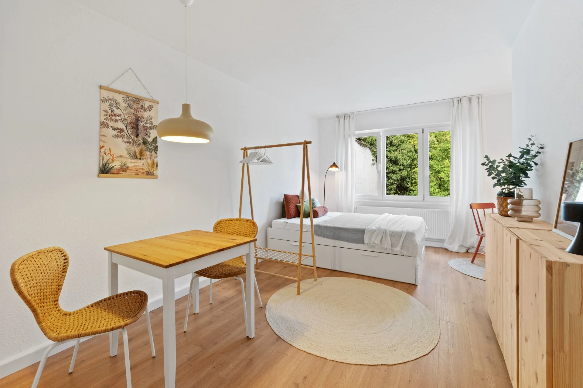 Helles Schlafzimmer mit Bett, Tisch, zwei Stühlen, Regal, rundem Teppich und großem Fenster.