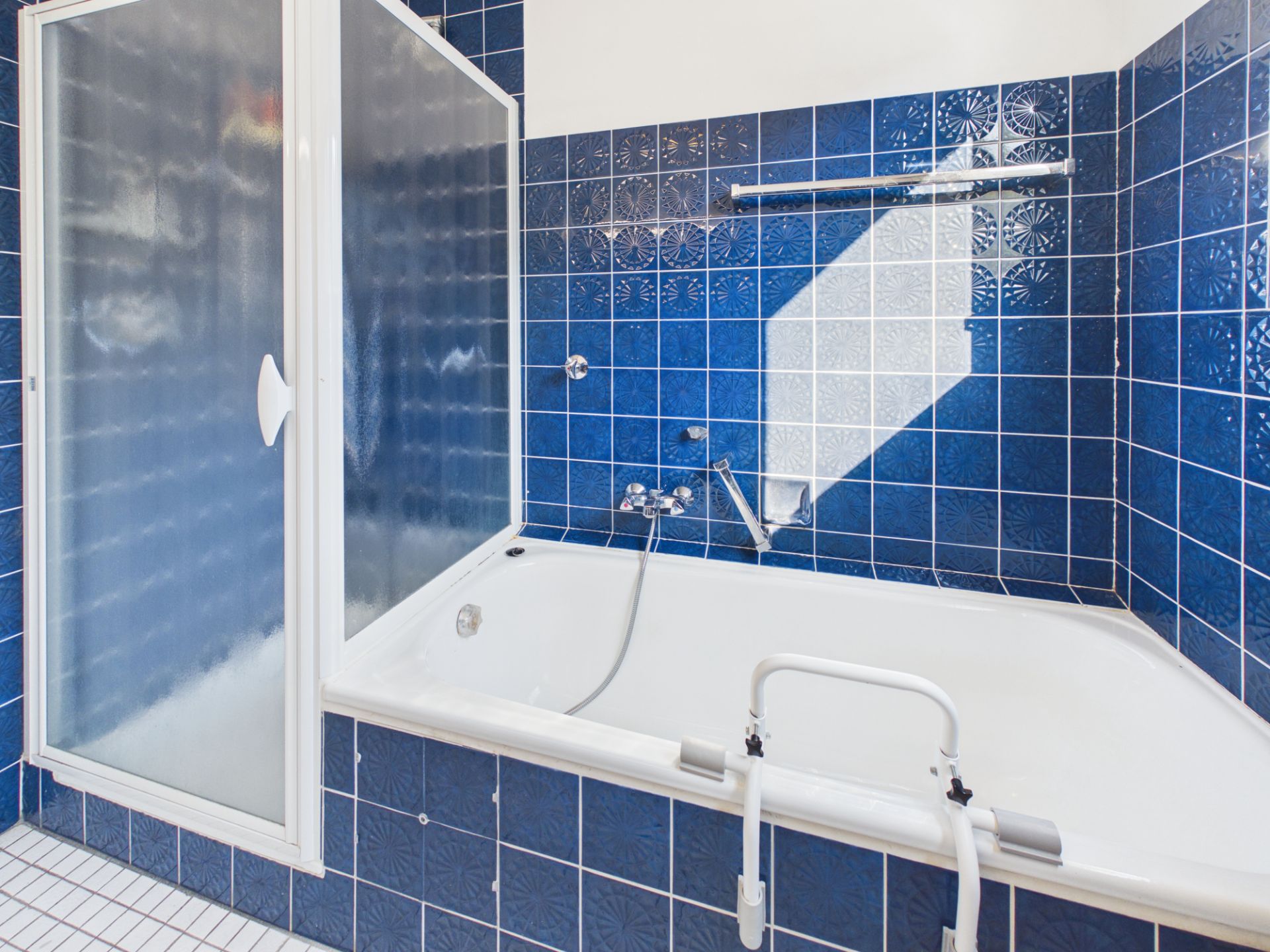 Badewanne mit Haltegriff und blauen Fliesen, daneben eine Dusche mit Glaswand.