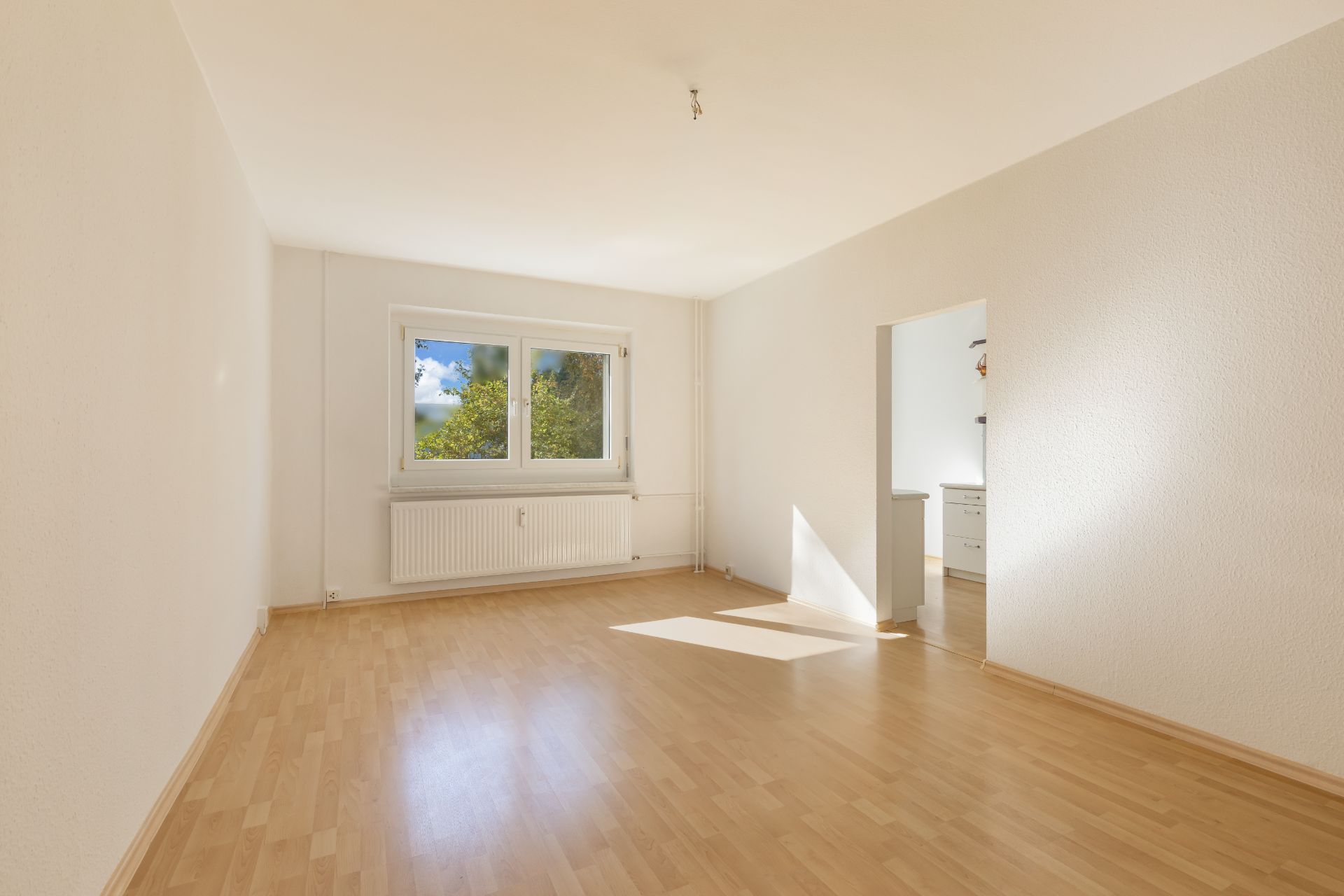 Helles, leeres Zimmer mit Holzfußboden und Fenster, angrenzender Raum sichtbar.