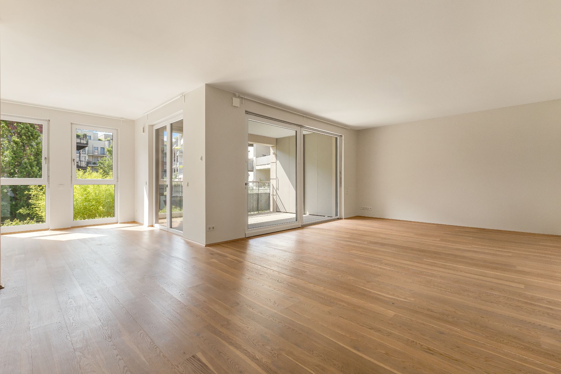 Helles, leeres Wohnzimmer mit Parkettboden und großen Fenstern zu einem Balkon.