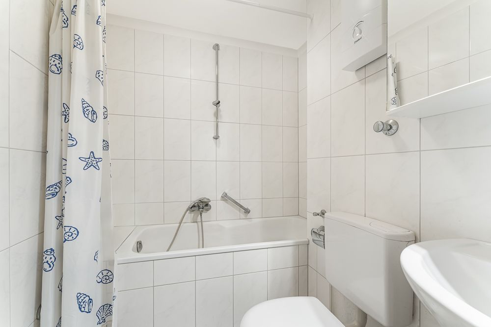 Weiß gefliestes Badezimmer mit Badewanne, Duschvorhang, Toilette und Waschbecken.