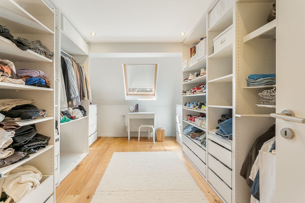 Offener, heller Ankleideraum mit Regalen, Kleidung, Schuhen und einem kleinen Schreibtisch unter einem Dachfenster.