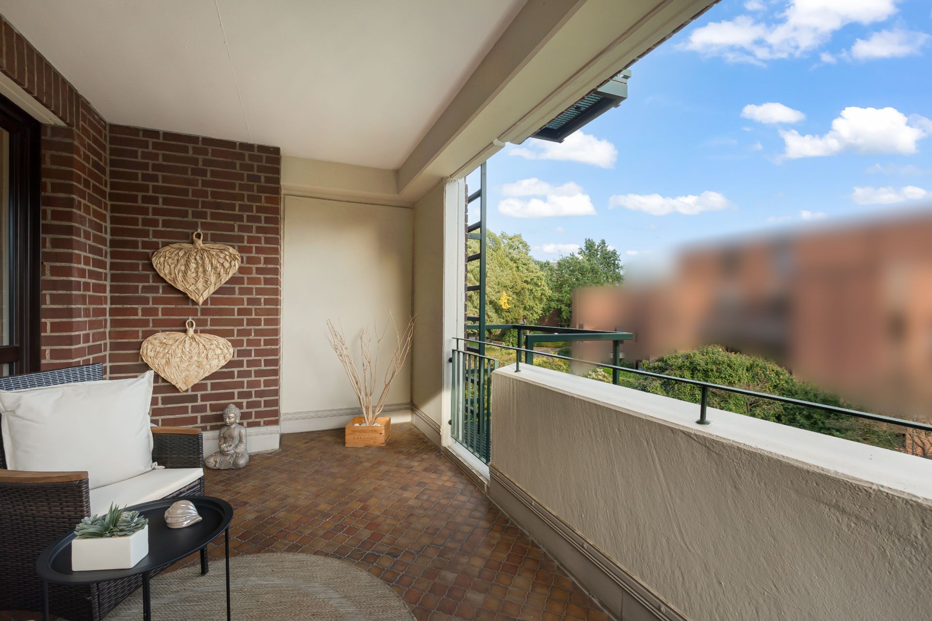 Überdachter Balkon mit Sitzgelegenheit, Deko und Blick auf Bäume und Gebäude.