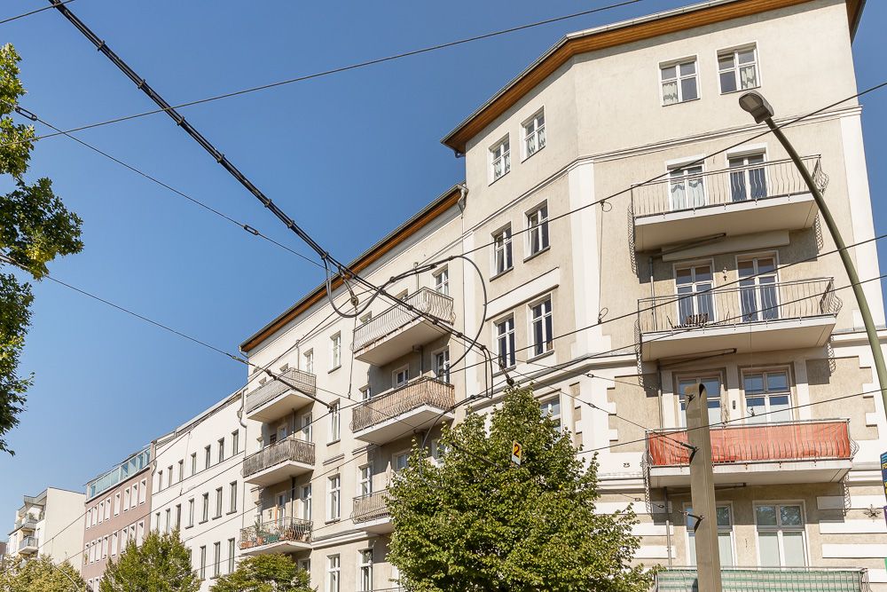 Mehrstöckiges Wohngebäude mit mehreren Balkonen, Bäumen davor und Stromleitungen im Vordergrund.