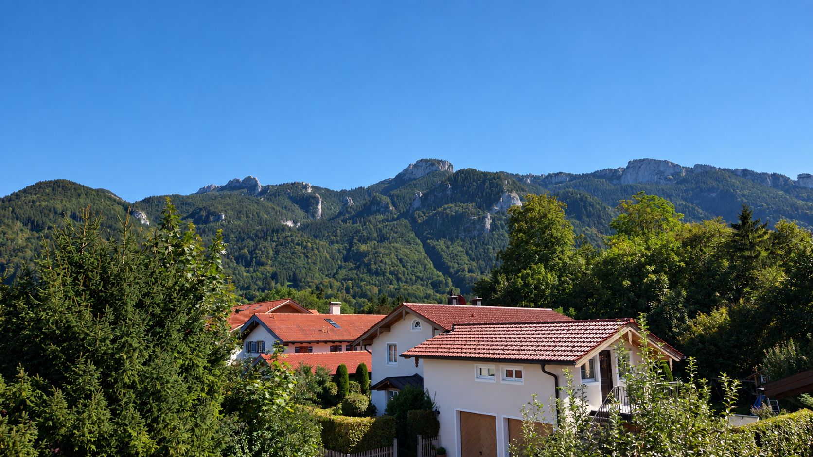 Häuser mit roten Dächern und grüner Gartenlandschaft vor bewaldeten Bergen unter blauem Himmel.