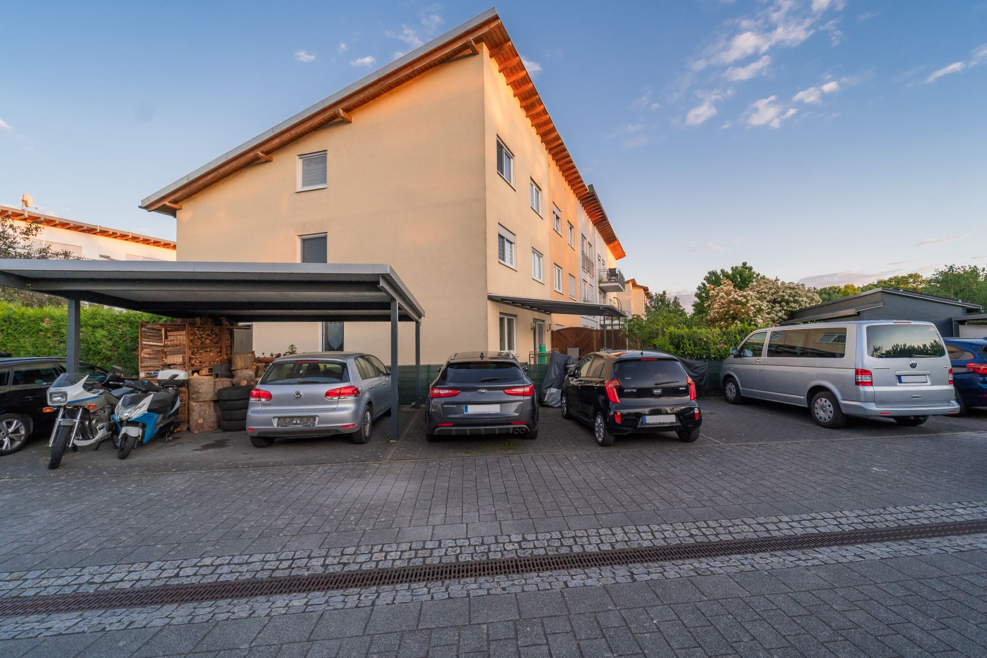 Mehrfamilienhaus mit Carport und geparkten Autos sowie Motorrädern auf einem gepflasterten Parkplatz.