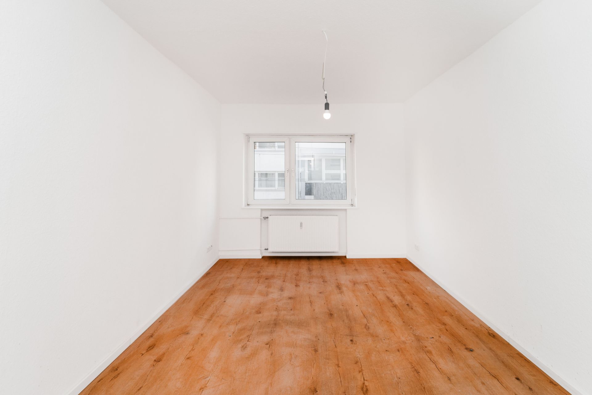 Leerer Raum mit weiß gestrichenen Wänden, Holzfußboden und einem Fenster mit Heizkörper.