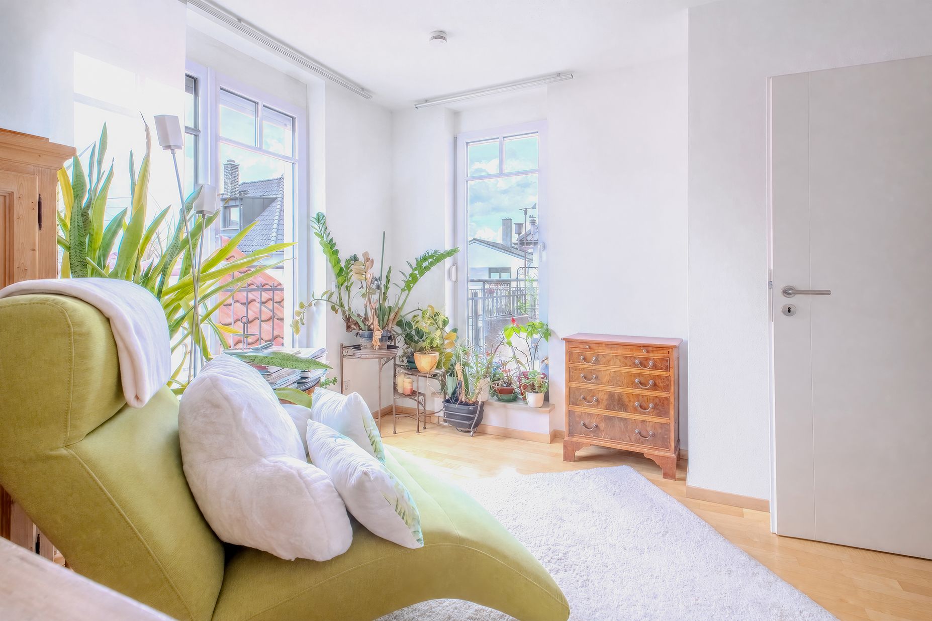 Helles Wohnzimmer mit grünem Sessel, vielen Pflanzen, Teppich und Kommode vor zwei Fenstern.