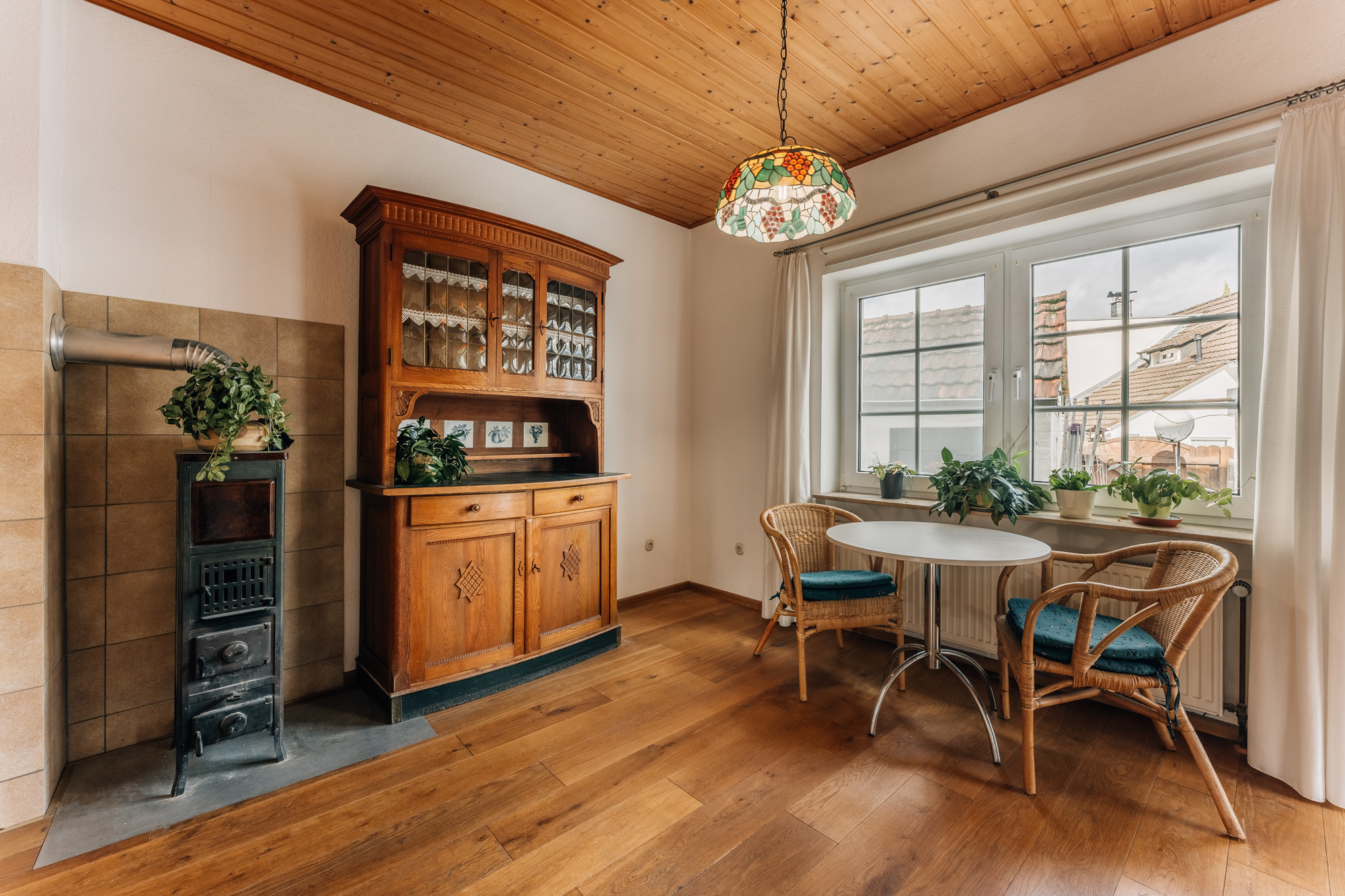 Essbereich mit rundem Tisch, zwei Stühlen, Holzschrank, Ofen und Pflanzen am Fenster.