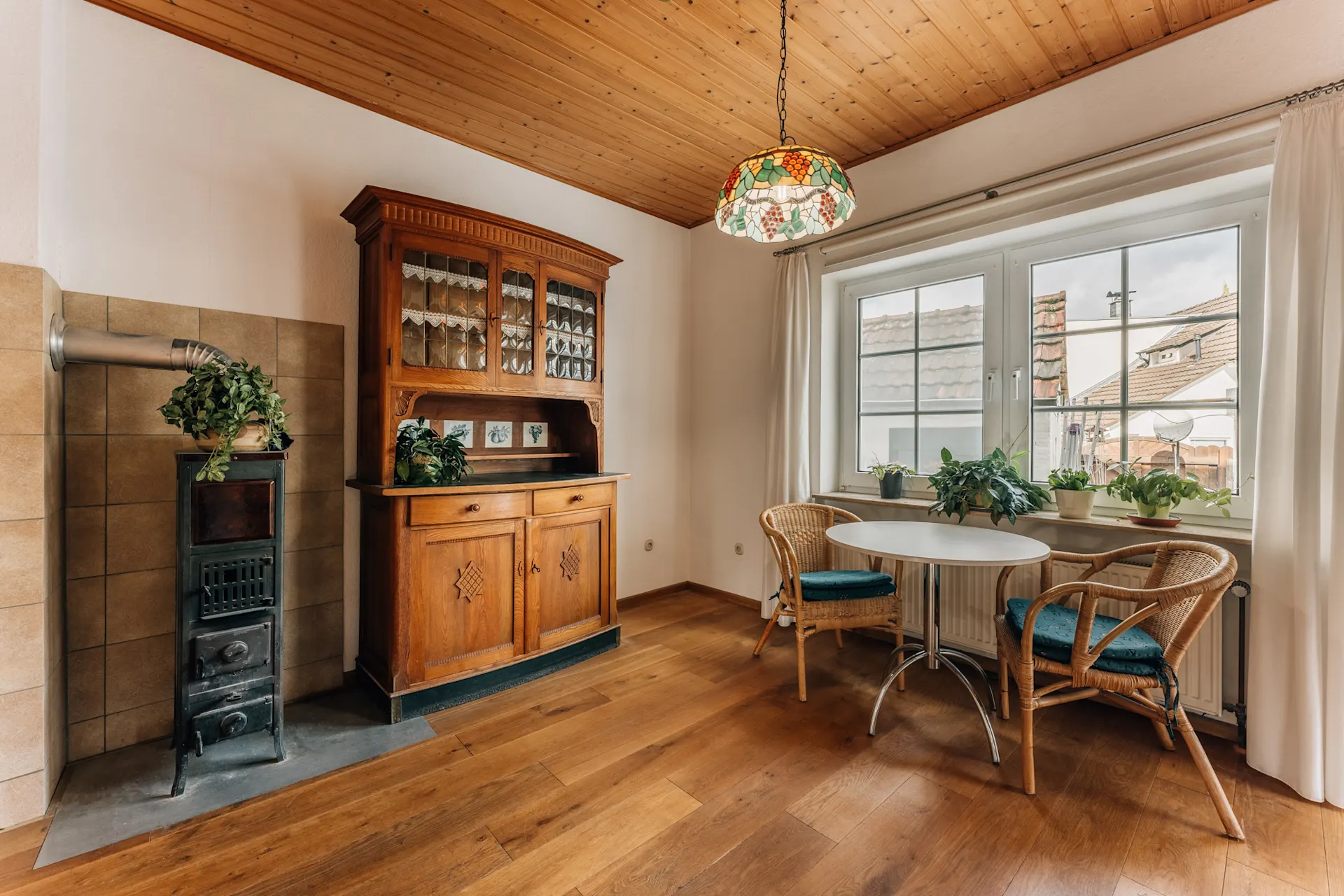 Essbereich mit rundem Tisch, zwei Stühlen, Holzschrank, Ofen und Pflanzen am Fenster.