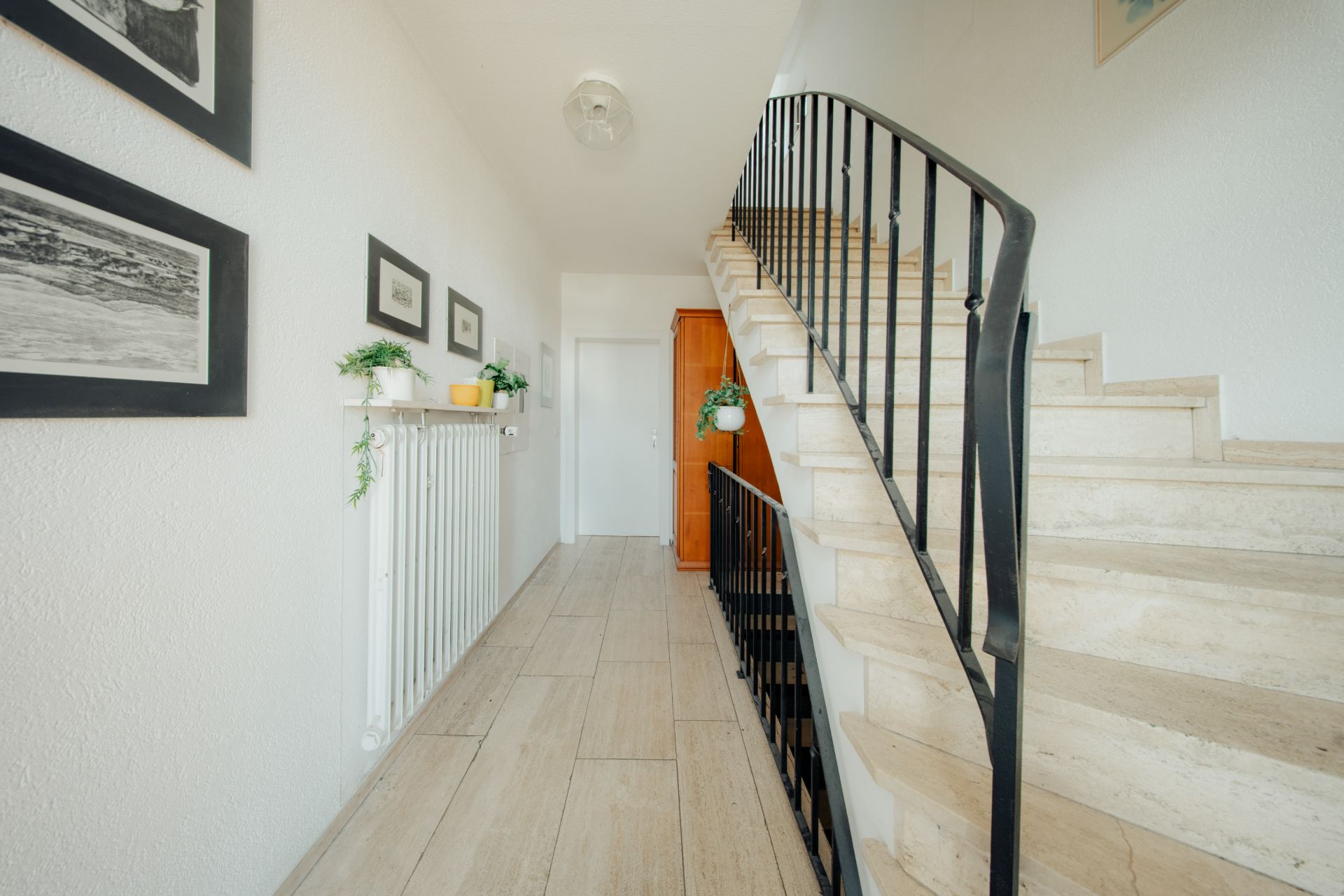 Heller Flur mit Treppe, an der Wand Bilder und Pflanzen, Holzschrank am Ende des Flurs.