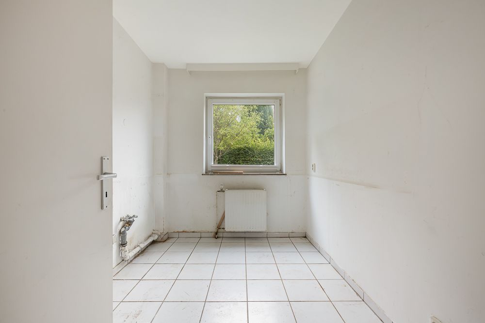 Leerer Raum mit weiß gefliesten Boden, einem Fenster und sichtbaren Rohrleitungen.