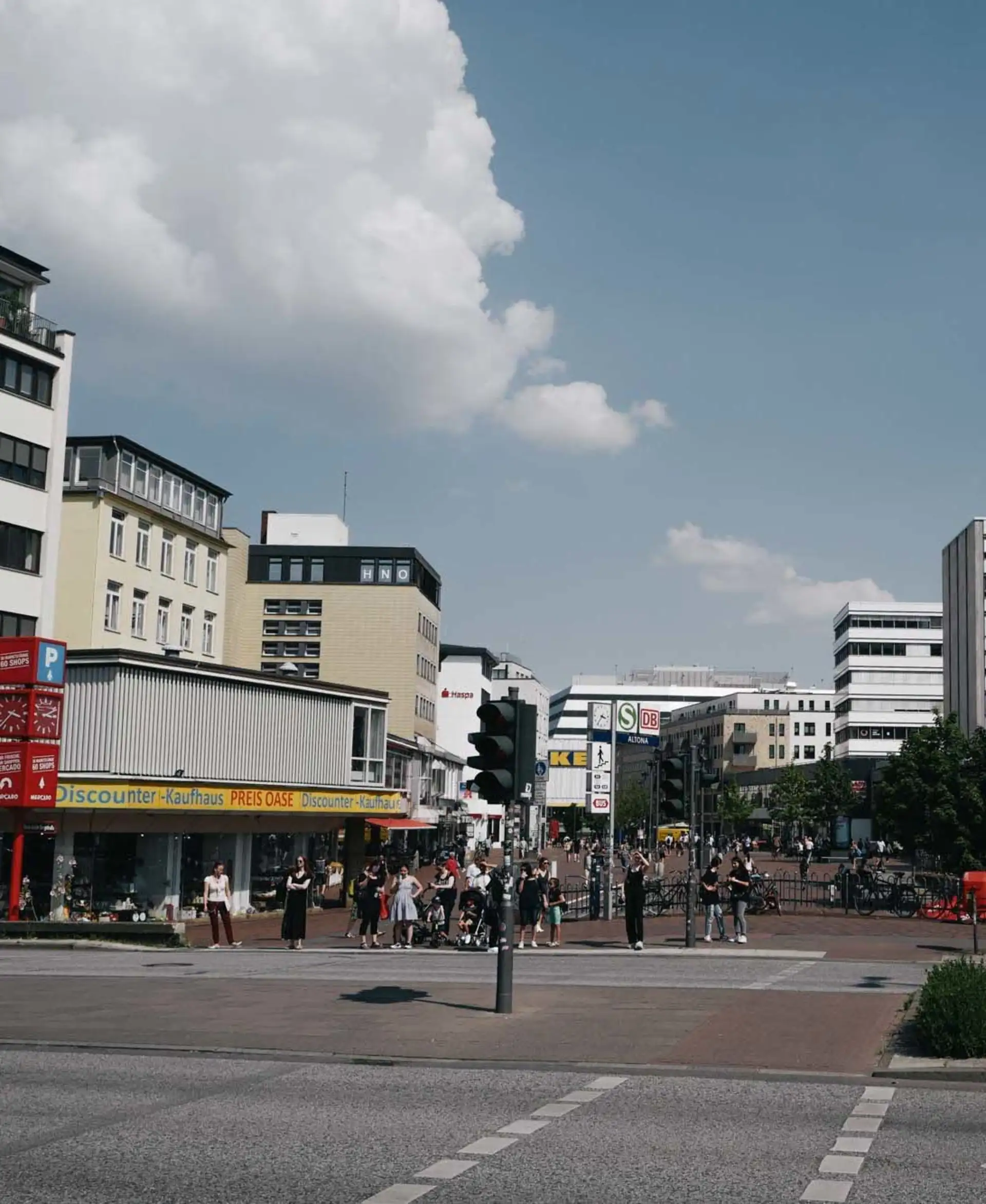 Straßenszene mit vielen Menschen, Geschäften und Ampeln an einer Kreuzung bei Tageslicht.