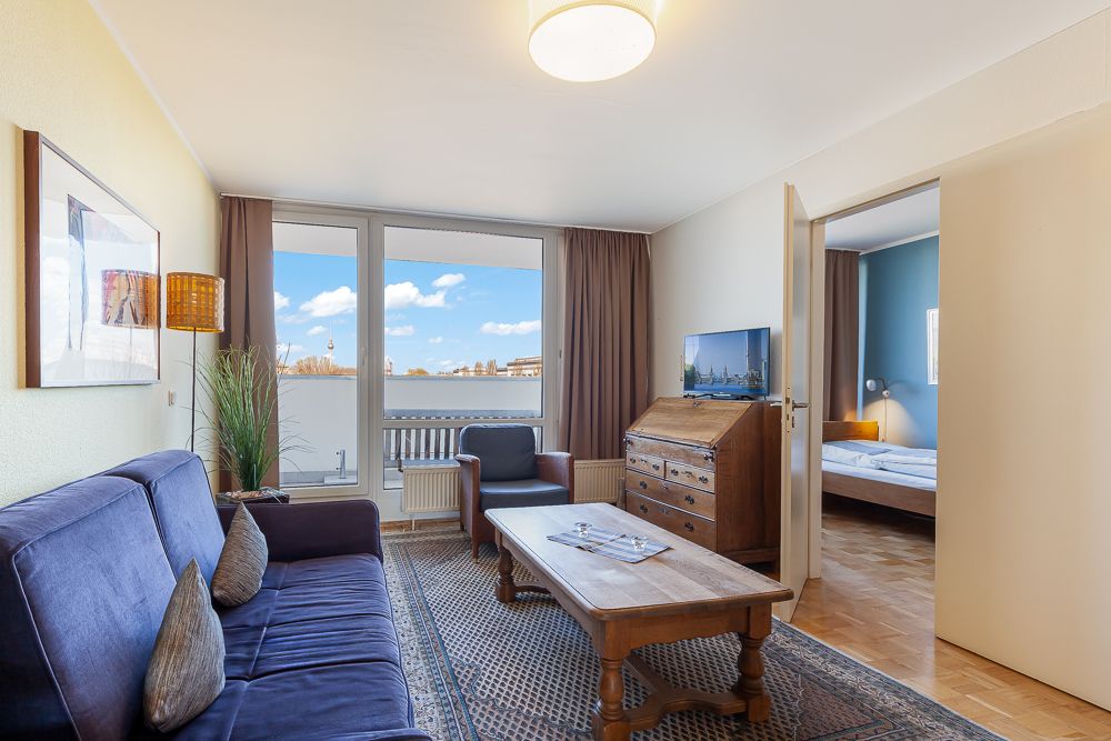 Wohnzimmer mit Sofa, Tisch, Sessel, Schreibtisch, Fernseher und Blick auf einen Balkon.